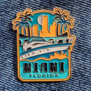 Puede incluir: Un pin rectangular de esmalte con un diseño de Miami, Florida. El pin muestra un yate, palmeras y un paisaje urbano sobre un fondo turquesa y naranja. La palabra "MIAMI" se muestra prominentemente, con "FLORIDA" debajo.