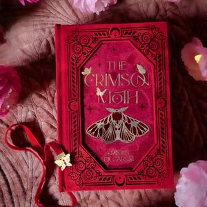 Puede incluir: Un libro rojo titulado "The Crimson Moth" con letras doradas y una ilustración de polilla. El libro tiene un marcapáginas de cinta roja con un dije de mariposa dorada. Flores rosas rodean el libro.