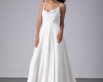 May include: A white wedding dress with a cowl neckline and spaghetti straps. The dress has a fitted bodice and a flowing A-line skirt. The fabric appears to be a lightweight linen or cotton, with a subtle texture. The dress is floor-length and has a simple, elegant design.