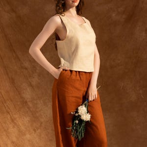 A woman wearing a beige top and brown pants stands against a brown background, holding a bouquet of white flowers.
