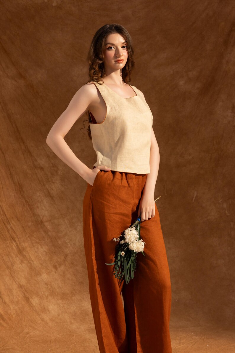 A woman wearing a beige top and brown pants stands against a brown background, holding a bouquet of white flowers.