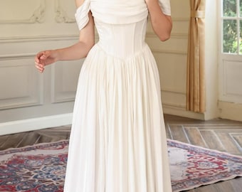 May include: A white off-the-shoulder wedding dress with a fitted bodice and flowing skirt. The dress features a high slit and is worn with white heels. The background includes a patterned rug and a window with natural light.