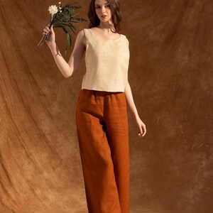 A woman in a white top and brown pants stands in front of a brown background, holding a bouquet of flowers.
