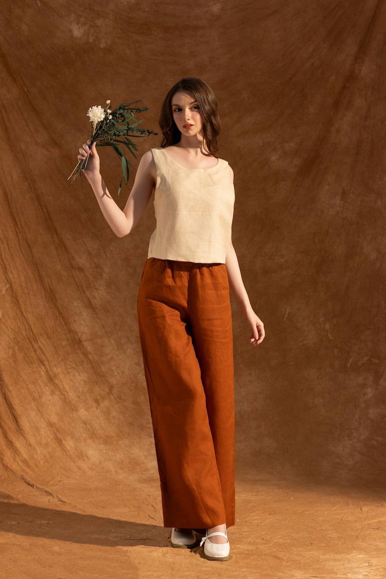 A woman in a white top and brown pants stands in front of a brown background, holding a bouquet of flowers.