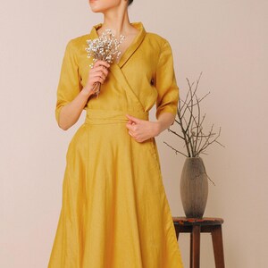 A woman in a yellow dress stands in front of a wooden table with a vase of flowers, holding a bouquet of white flowers.