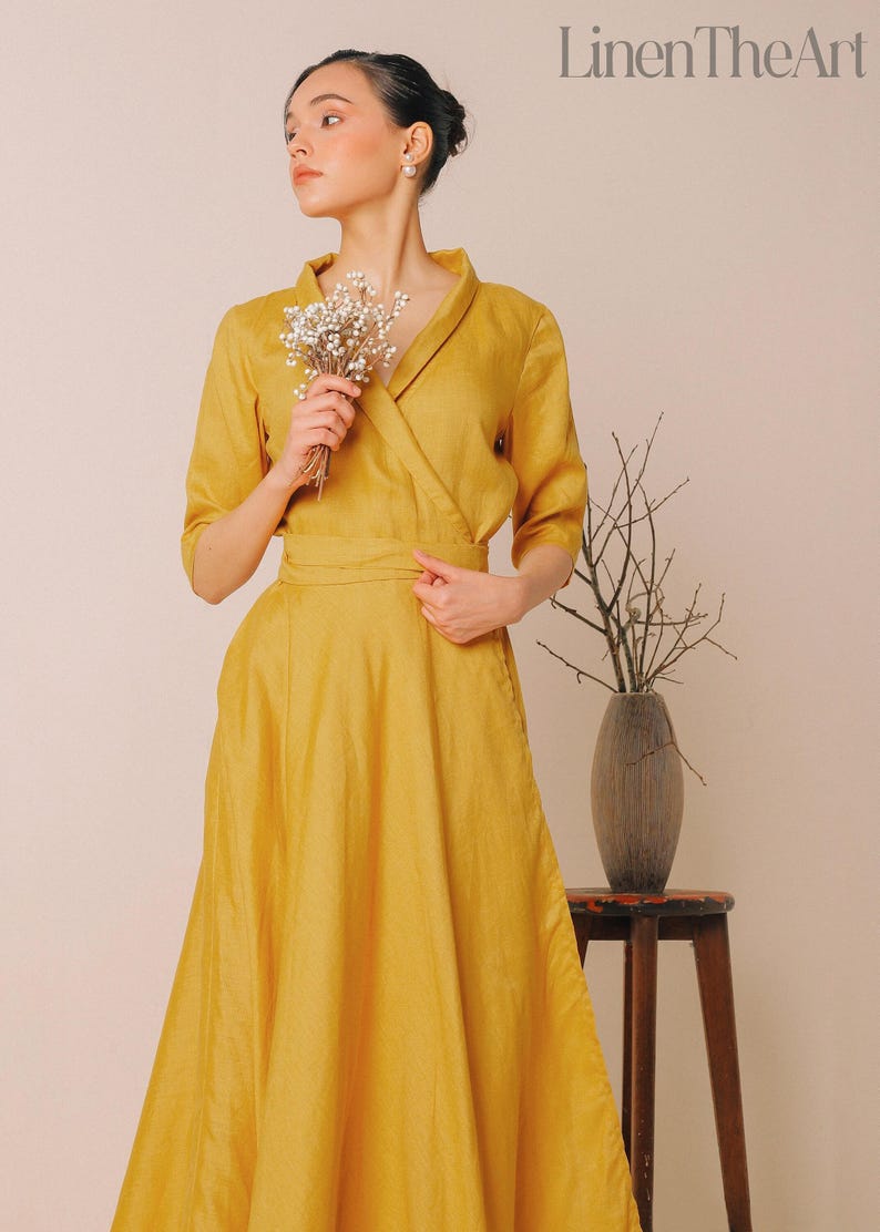 A woman in a yellow dress stands in front of a wooden table with a vase of flowers, holding a bouquet of white flowers.