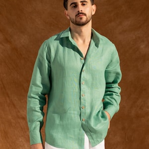 May include: A light green button-down shirt with long sleeves. The shirt is made of a textured fabric and has a classic collar. The model is wearing white pants. The background is a solid brown color.