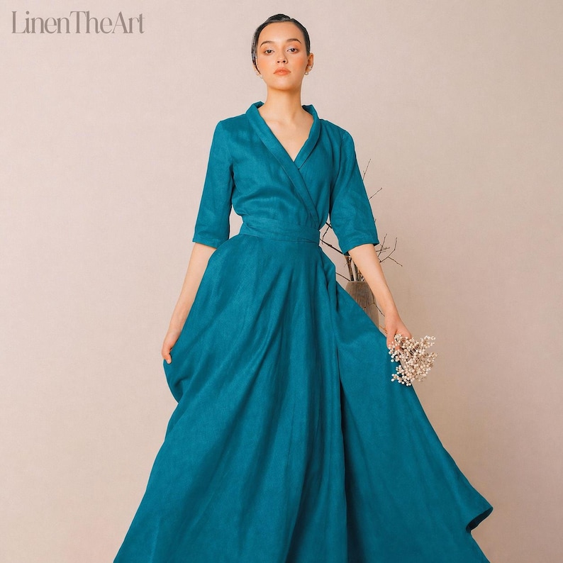 A woman wearing a long, flowing teal dress stands against a light background, holding a small bouquet of flowers.
