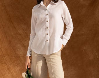 May include: A white long-sleeve button-down shirt with wooden buttons and a collared neckline. The shirt is paired with beige wide-leg pants. The person is holding a small bouquet of white flowers and greenery.