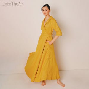 A woman wearing a long, yellow dress with a wrap-style neckline and a high collar, posing against a plain white background.