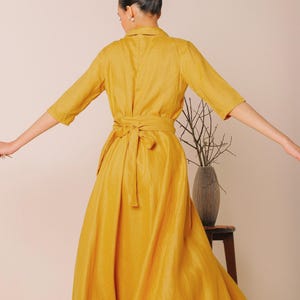A woman in a long yellow dress stands with her arms outstretched, facing away from the camera.