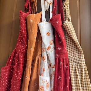 May include: A selection of colourful aprons hanging on a door. The aprons are in various shades, including red with white polka dots, orange, white with floral patterns, and red with bow accents. A brown and white checked apron is also visible.