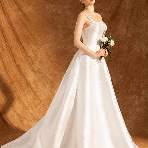 May include: A white satin wedding gown with a square neckline and spaghetti straps. The dress has a full skirt and a long train. The person is holding a small bouquet of white flowers. A thin, white headband is worn.