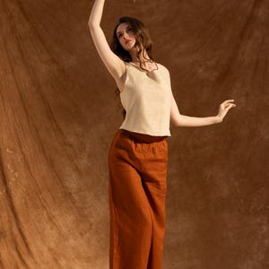 A woman in a beige top and brown pants stands in front of a brown background, holding a bouquet of flowers in her hand.