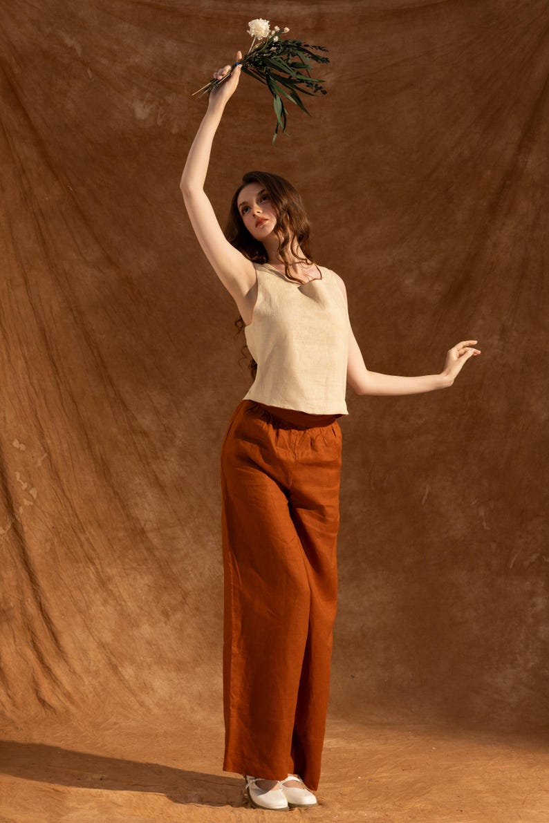A woman in a beige top and brown pants stands in front of a brown background, holding a bouquet of flowers in her hand.