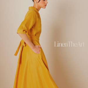 A woman wearing a long, yellow dress with a high collar and a tie at the waist, posing against a plain background.