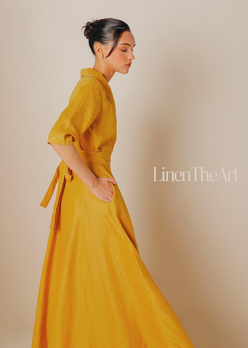 A woman wearing a long, yellow dress with a high collar and a tie at the waist, posing against a plain background.
