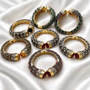Pode incluir: Uma coleção de sete pulseiras abertas com armações de metal dourado e detalhes pretos intrincados. Cada pulseira é adornada com várias pedras preciosas ovais coloridas, incluindo vermelho, verde e amarelo, acentuadas por pequenos cristais transparentes.