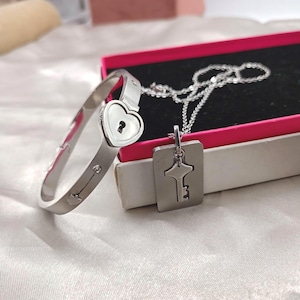 Puede incluir: Una pulsera plateada con un diseño de cerradura en forma de corazón y un ojo de cerradura, junto a un collar a juego con un colgante de llave. Las joyas se exhiben cerca de una caja rosa y negra, lo que sugiere un juego de regalo.