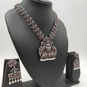 May include: A silver-toned necklace and earring set displayed on a black mannequin. The necklace features a pendant with a seated figure and pink accents. The earrings mirror the necklace design, with a floral pattern and dangling pearls.