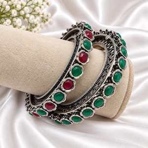 May include: Two silver-toned metal bangles featuring an intricate filigree design. Each bangle is adorned with a repeating pattern of circular, bezel-set cabochon stones in alternating vibrant green and deep red colors along the outer edge.