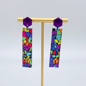 May include: A pair of colorful, geometric patterned earrings with purple hexagon-shaped tops. The earrings are displayed on a gold stand.