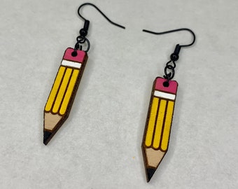 Puede incluir: Un par de pendientes de lápiz amarillo con gomas de borrar rosas y ganchos de metal negro. Los pendientes están hechos de madera y tienen un diseño de lápiz realista.