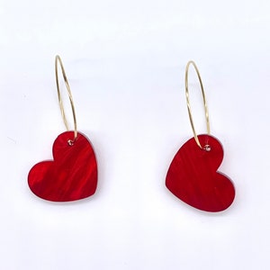 May include: A pair of gold hoop earrings with red heart-shaped charms. The hearts have a marbled, textured look.