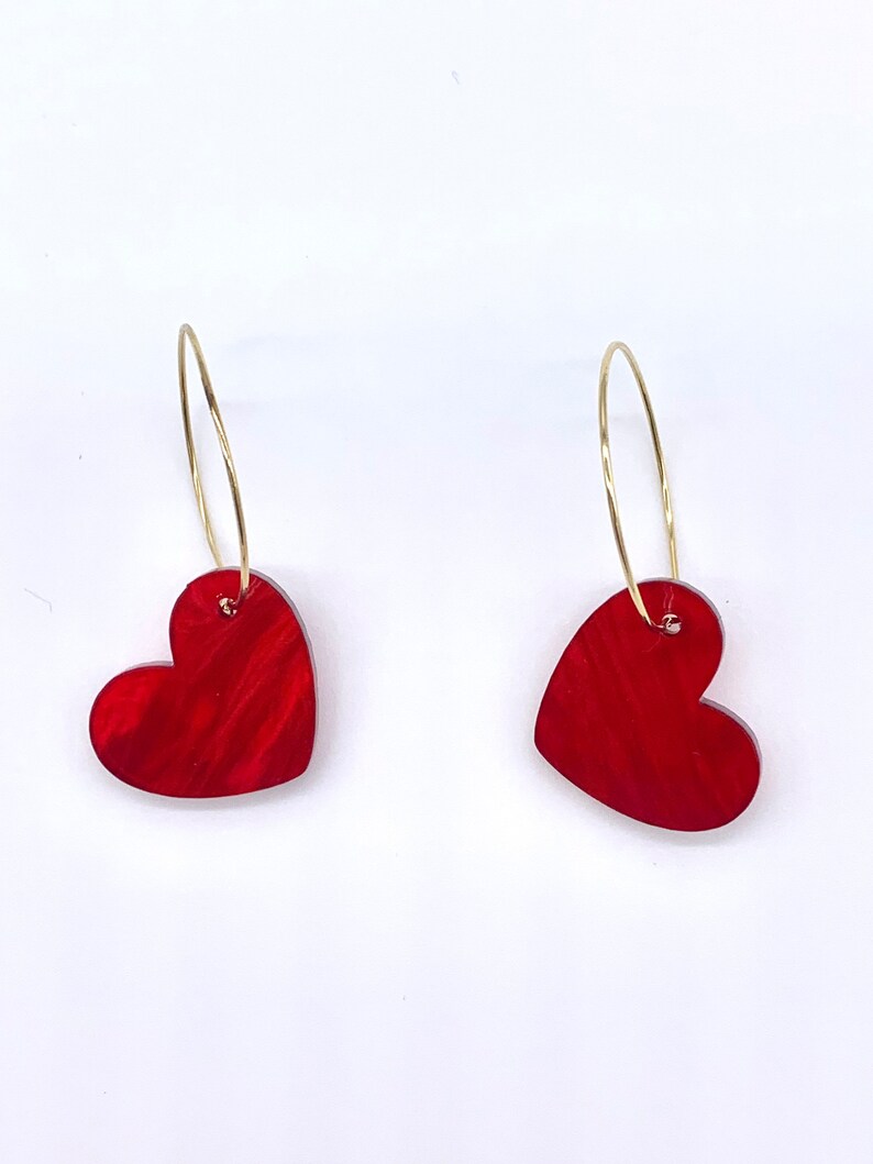 May include: A pair of gold hoop earrings with red heart-shaped charms. The hearts have a marbled, textured look.
