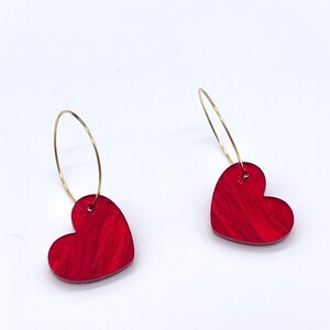 May include: A pair of gold hoop earrings with red heart-shaped charms. The hearts have a marbled pattern.