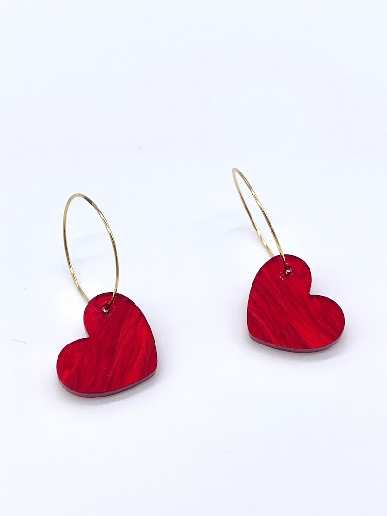 May include: A pair of gold hoop earrings with red heart-shaped charms. The hearts have a marbled pattern.