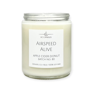 May include: A white candle in a clear glass jar with a black label that reads "XC CANDLES", "AIRSPEED ALIVE", "APPLE CIDER DONUT", "BATCH NO: 80", and "DENVER CO / 8OZ / 100% SOY WAX".