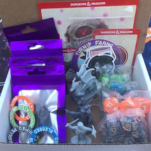 May include: A box filled with Dungeons & Dragons themed items, including dice, a dragon miniature, and a Turnip Farms sticker. There are also several purple bags with silver rings inside.