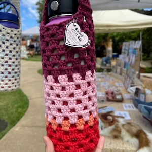 Puede incluir: Una bolsa de ganchillo hecha a mano para botella de agua en tonos de rojo, rosa y burdeos. La bolsa presenta un diseño de granny square y una pequeña etiqueta que dice "Crochet Bottle Bag". La bolsa está diseñada para contener una botella de agua.
