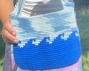 May include: A blue and white crochet shoulder bag. The bag features a gradient design, transitioning from light blue to a deep blue at the bottom. The bag has a wave-like pattern near the bottom and a matching strap.