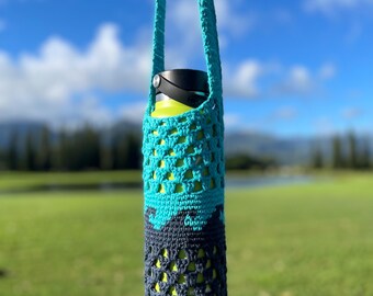 May include: A teal and navy blue crocheted water bottle holder with a matching strap. The holder is designed with an open weave pattern, allowing the yellow water bottle inside to be partially visible. The background features a green field and a blue sky.
