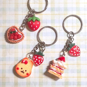 May include: Keychains with food-themed charms. The charms include a red strawberry, a heart-shaped tart with red berries, a smiling pear-shaped cookie with a red bow, and a layered pastry. Each charm is attached to a silver keyring.