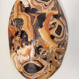 May include: An oval-shaped decorative stone featuring a marbled pattern with swirling colors of dark brown, tan, cream, black, and burnt orange. The surface displays a glossy finish with distinct layered textures and organic, fluid shapes throughout.