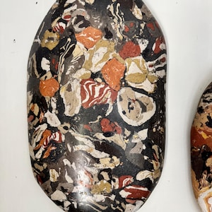 May include: An oval-shaped decorative stone featuring a complex, multi-colored marbled pattern. The surface displays a dense mix of black, white, tan, orange, and deep red swirls and irregular patches, creating a highly textured, variegated appearance.