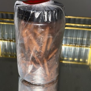 Puede incluir: Un tarro transparente envuelto en plástico con tapa negra, que contiene palitos marrones. El tarro está sobre una superficie reflectante con borde dorado. La parte superior del tarro tiene una etiqueta roja y blanca.