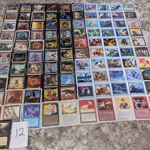 May include: A large collection of rectangular trading cards arranged in a grid pattern, featuring diverse colorful illustrations of creatures, landscapes, and magical symbols. A small white card with the handwritten number 12 is placed at the bottom.