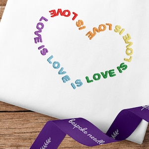 Op de afbeelding: Witte stof met een regenboog hart geborduurd met de tekst "Love is Love" die herhaald wordt rond het hart.