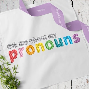 May include: White tote bag with the embroidered text "ask me about my pronouns" in a rainbow of colors. A lavender ribbon with the text "bespoke needle" is draped across the bag. Greenery is visible in the lower left corner.