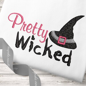 May include: Embroidered design on a white t-shirt featuring the text "Pretty Wicked" in black and pink with a black witch hat with a pink buckle.