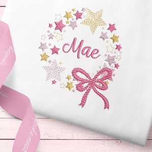 May include: A pink and gold embroidered wreath with stars and a bow. The wreath says "Mae" in the center.