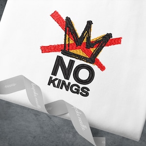 May include: White fabric with an embroidered design featuring a black and yellow crown crossed out with a red line. The text "NO KINGS" is embroidered below the crown in black. A gray ribbon is in the foreground.