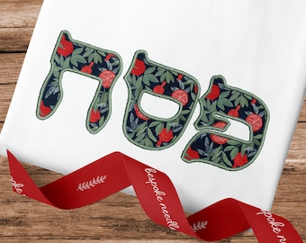 May include: White fabric with the Hebrew word "Shabbat" embroidered in green and red. The letters are filled with a pattern of pomegranates and leaves.