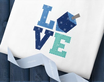 May include: A white t-shirt with the word "LOVE" spelled out in blue and teal fabric. The "O" is replaced with a blue and white dreidel.