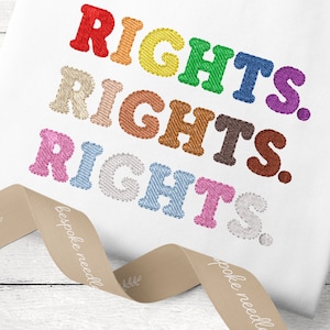 Op de afbeelding: Een wit T-shirt met het woord "RIGHTS" geborduurd in regenboogkleuren. Het woord wordt drie keer herhaald, waarbij elke herhaling in een andere kleur is.