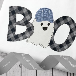 May include: White onesie with a black and white plaid "BOO" applique. The "O" is a ghost wearing a blue baseball cap.